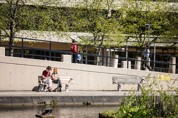 Two students sit on a bench next to a pond on the University of Leeds campus looking at an electronic device