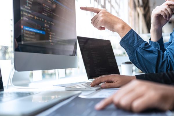 Two people point at computer screens on a desk