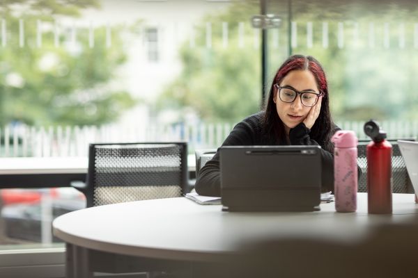 Students sit at a round table in a bright campus study area, working on tablets and a laptop while taking notes.