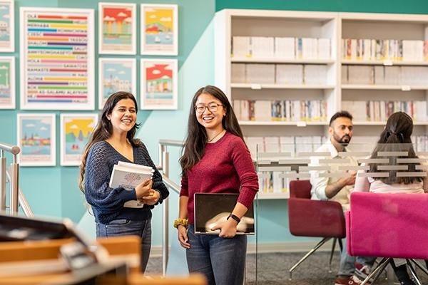 Two students holding books stood together in the Language Centre smiling. There are colourful photos on the blue wall in the background.