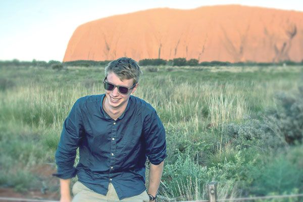 A student leans on a railing, smiling. Behind them is grasslands and a large rust-coloured mountain.