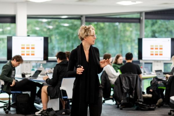 An academic tutor stood teaching in the middle of a large, busy classroom. The modern room has big, bright windows along the side, with large digital screens and clusters of people working together on their laptops, around shared desks.