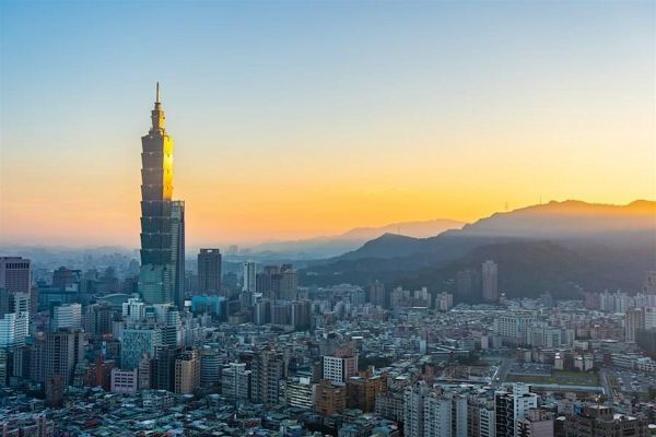 Skyline of Taipei at sunset. Large skyscrapers soar over buildings with mountainous landscape in the background.