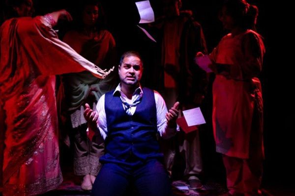 An actor on stage gesturing with their hands, lit by a dramatic spotlight, and surrounded by a few other actors.