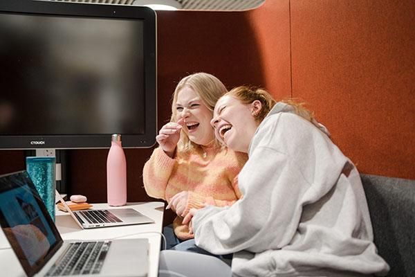 Two students lean against each other and laugh whilst working on their laptops inside a booth