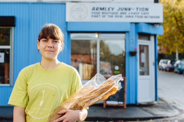 A smiling student volunteer stands holding baguettes in front of Armley Junk-tion cafe.