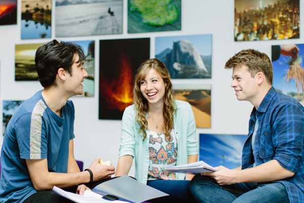 Three students sat around a table talking with landscape photography on the wall behind them.