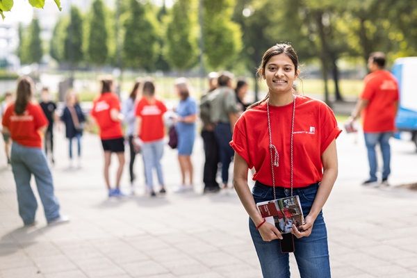一位开放日大使穿着红色t恤站在校园里对着镜头微笑。背景是穿着红色t恤的开放日与会者和其他开放日大使