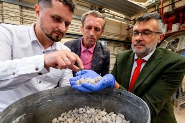 Three people in a workshop inspect a bucket of small stones; one holds some stones in gloved hands while the others observe.