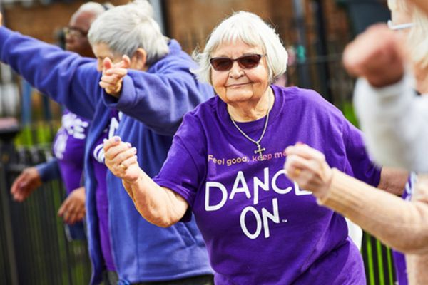 An elderly person dancing in a group, wearing a 'Dance On' t-shirt and sunglasses.