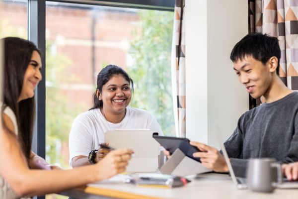 Three students sit at a dining table in residential halls on campus. Two are holding tablets and the third is holding a pen. They are all smiling.