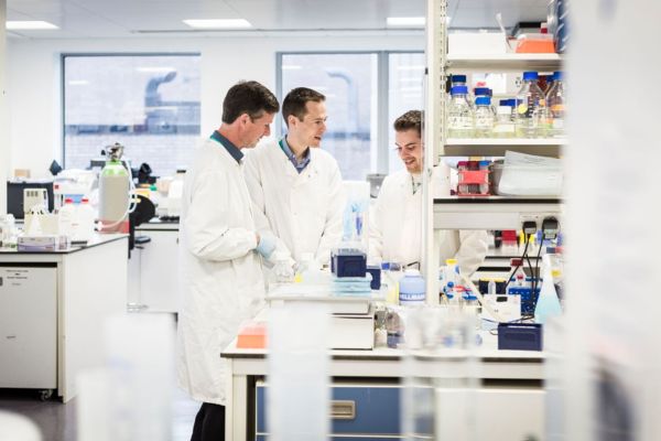 Three researchers work together in a laboratory. They are all wearing white lab coats and chatting. Several benches with various scientific equipment surrounds them.