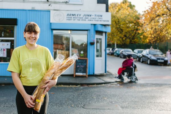 A smiling student volunteer stands holding baguettes in front of Armley Junk-tion cafe with a person in a wheelchair going up a kerb in the background.