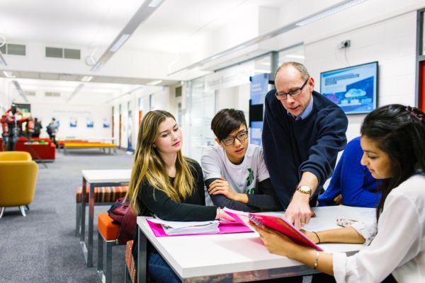 A teacher helping four students sat at a table, pointing at something in a book.