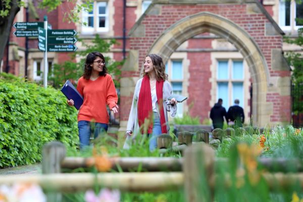 Two students walk across campus, away from a redbrick archway and buildings, along a path lined with greenery.