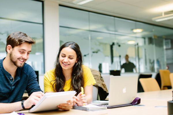 Two Masters students sat together in the library, looking at a notepad and smiling.