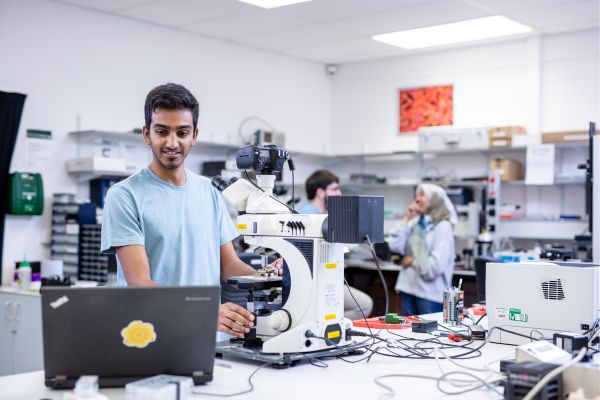 A researcher working with a machine in a lab, with colleagues chatting nearby.