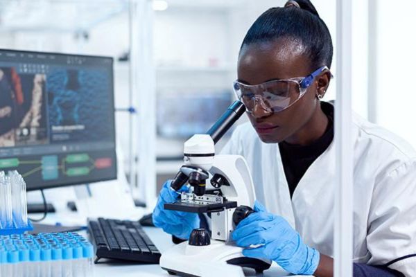 A researcher in a lab coat and goggles sat at a desk looking through a microscope