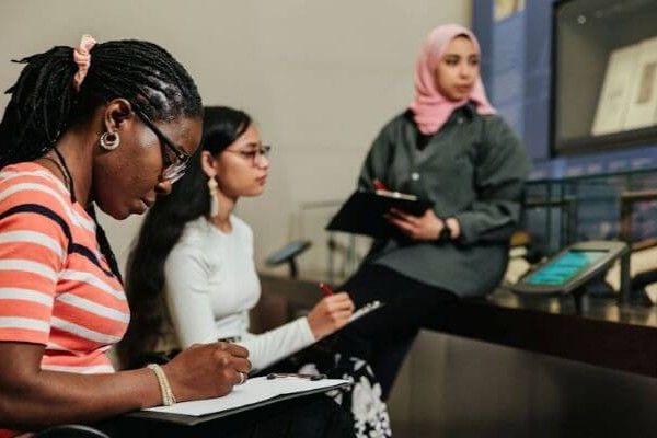 Three women sit writing on notepads, looking at a museum exhibit.