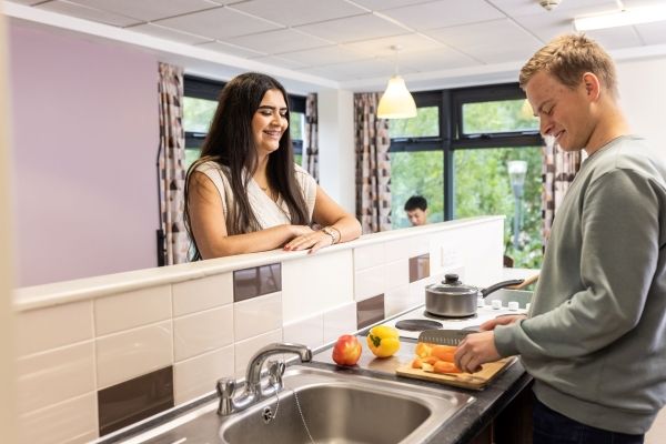 Two student stand chatting in a kitchen in residential halls. One is preparing food next to a sink, the other is leaning on the counter that acts as a backsplash. There are two other students sat at a table in the background.