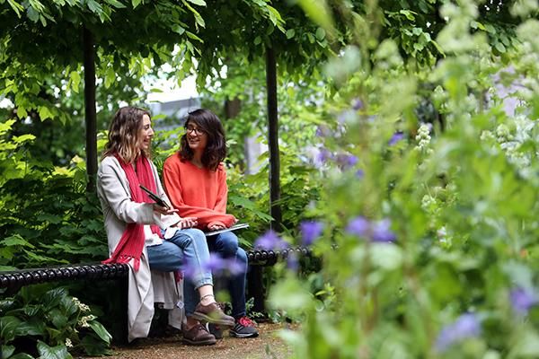 Two students sitting on a bench surrounded by trees and purple flowers.