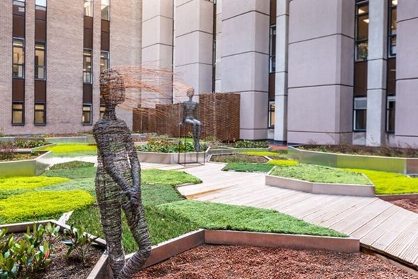 A green space with two wire sculptures of human figures, Buildings are in the background.
