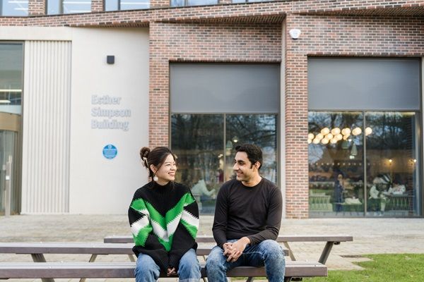 Two students chatting while sat on a bench outside the Esther Simpson Building.