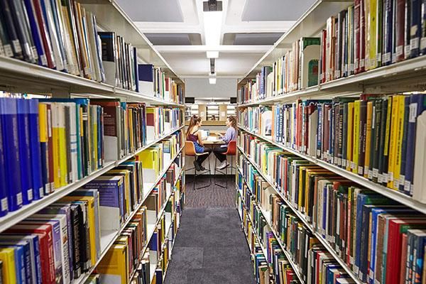 Two rows of books in a library with two students working in the distance.