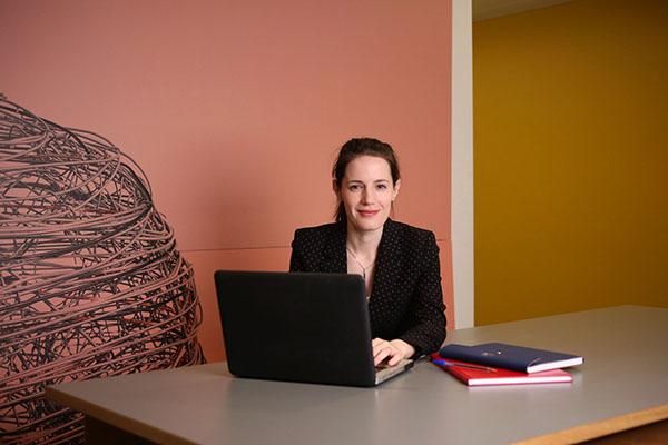 A person in a black jacket sits at a table with a laptop and books in front of them.