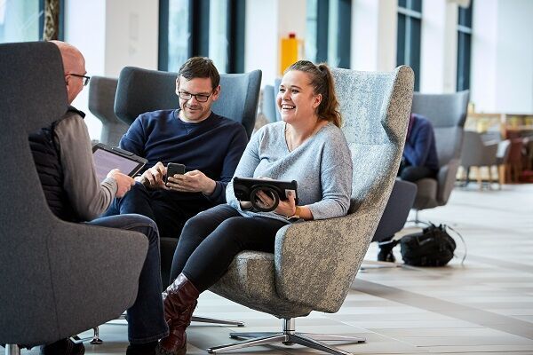 Three people sat in big comfy chairs in a study space, chatting and smiling. One is looking at a mobile phone, the other two are using tablets