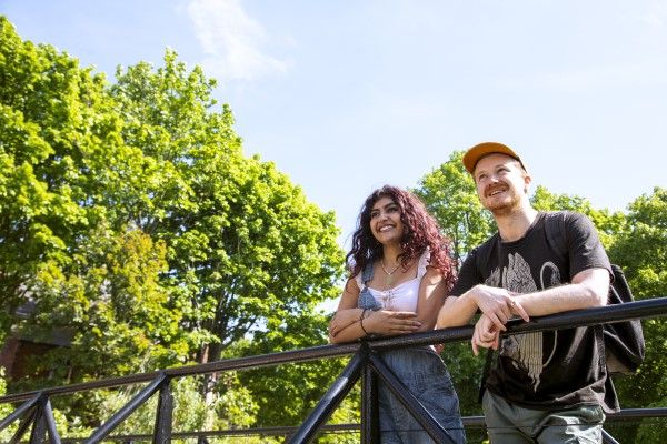 Two smiling students stood on a balcony outside on campus with trees in the background.