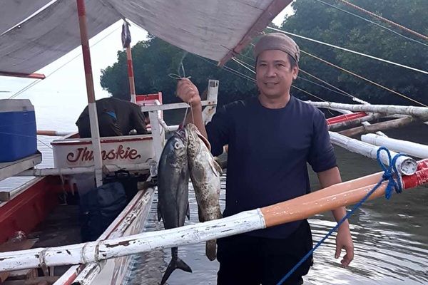 A person stood next to a boat in shallow waters, holding up two large fish they have caught.
