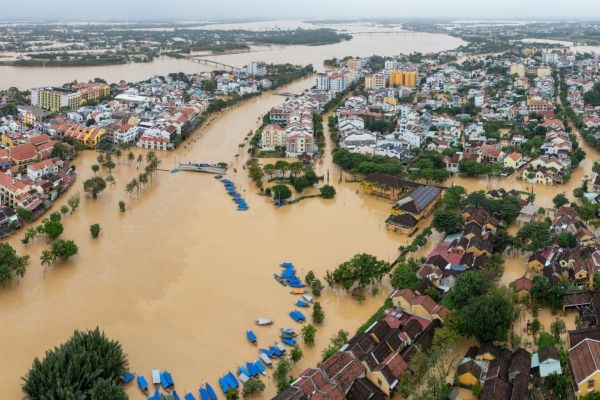 Aerial view of a densely built riverside town with brown floodwaters submerging streets, surrounding houses, and trees, with boats navigating through the inundated area.