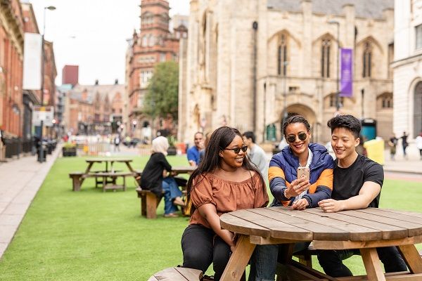Three students sat on a picnic bench in Leeds city centre. One is holding a mobile phone which they are all looking at and smiling.
