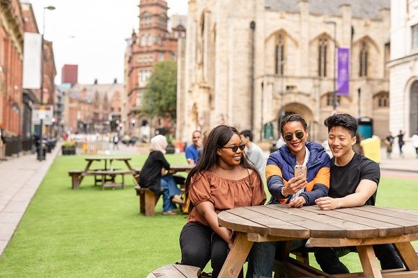 Three students sat on a picnic bench in Leeds city centre. One is holding a mobile phone, which they are all looking at and smiling.