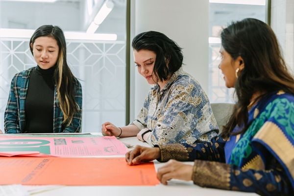 Three people sat at a large desk, discussing some bright printed materials spread out in front of them.