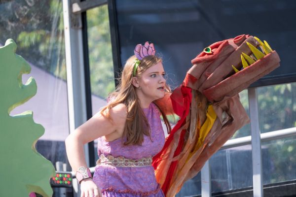 An actor wearing a purple dress and flower headband on stage, holding a dragon puppet with their left hand.