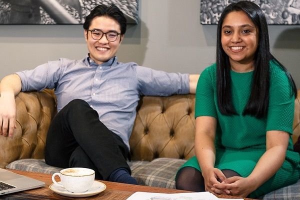 Two students sit on a sofa in a cafe, smiling. In front of themthere is a coffee table with a cup of coffee, laptop and documents on it.