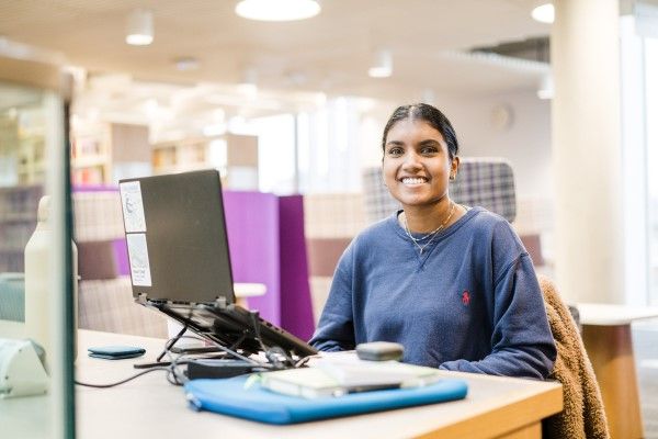 A student sits and smiles towards the camera while working on a laptop inside a library.