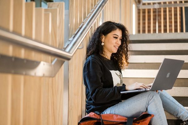 A student is sat on some steps in a modern interior space on campus, looking at a laptop and smiling.