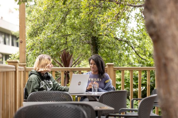 Two students sit at a small outdoor table on wooden decking using a laptop.
