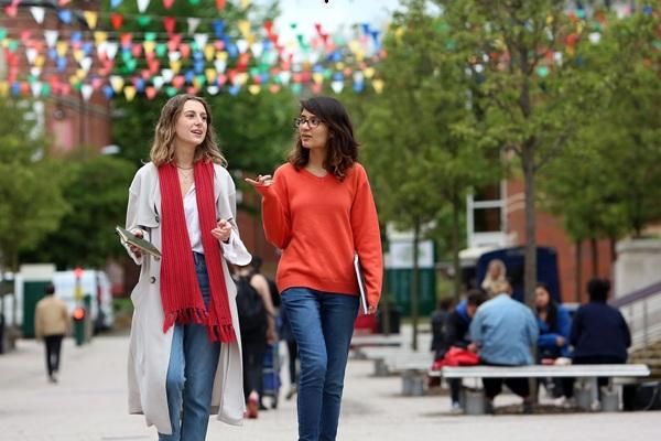 Two students walking towards the camera chatting and gesturing with their hands outside the LUU building.