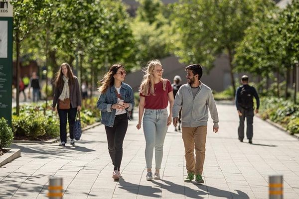 Three students walking on campus on a sunny day, chatting. There are lots of green trees and some other students in the background.