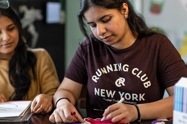 Two students seated at a table taking part in a creative craft activity, with hands working on colourful materials in a shared community workspace.
