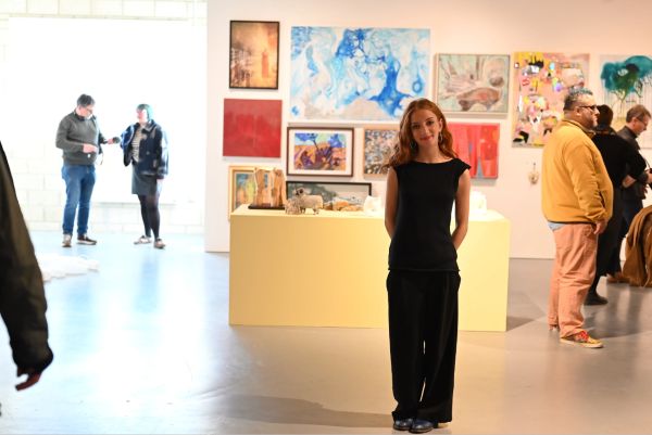 A person standing in an art gallery, with colourful artworks displayed on the walls and visitors in the background.