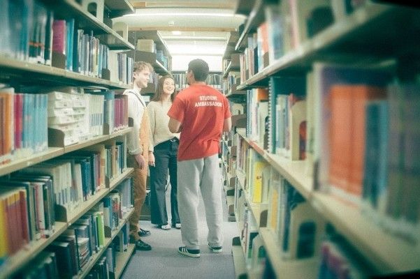 Two students stand talking to a student ambassador who is turned away from the camera. They are at the end of an aisle of books in a library.