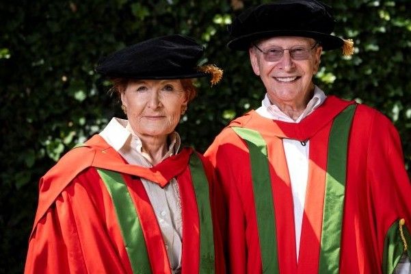 Peter and Susan Cheney smiling at the camera in red and green graduation robes with black caps.