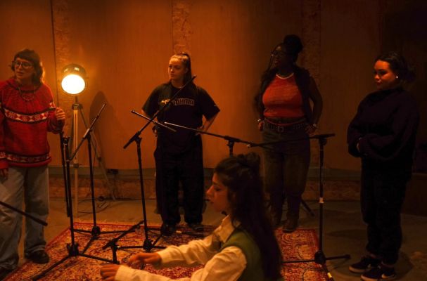A group of five individuals gathered in a studio space with microphones set up, illuminated by warm lighting, as one person sits on a rug in the centre.