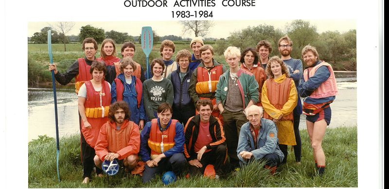 A group of students gather before a river, some wearing life jackets and other holding oars