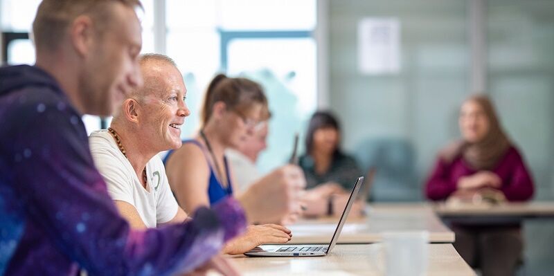 A group of smiling mature students sat in a classroom listening to someone speaking off camera